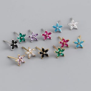 Peut inclure: Une collection de boucles d'oreilles à clous en forme de fleurs de différentes couleurs, dont le noir, le vert, le violet, le rose, le blanc et le turquoise. Certaines boucles d'oreilles ont des tiges dorées, d'autres des tiges argentées.