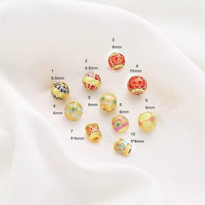 Peut inclure: Dix perles dorées avec des motifs en émail. Les perles sont de différentes tailles et couleurs, notamment rouge, bleu, vert, rose et blanc. Les perles sont disposées en cercle sur une surface blanche.