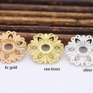 Puede incluir: Tres adornos metálicos decorativos con forma de flor con un agujero en el centro. Los adornos están en tres acabados diferentes: oro, latón y plata. El texto "kc gold", "raw brass" y "silver" está impreso debajo de cada adorno.