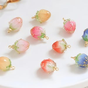Puede incluir: Una colección de coloridos colgantes de capullos florales en varios tonos de rosa, naranja, rojo y azul. Cada colgante tiene un lazo dorado para sujetarlo a la joyería. Los colgantes están dispuestos sobre una superficie blanca.