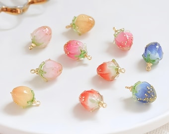 Può includere: Una collezione di ciondoli colorati a forma di boccioli di fiori in varie tonalità di rosa, arancione, rosso e blu. Ogni ciondolo ha un anello color oro per il fissaggio ai gioielli. I ciondoli sono disposti su una superficie bianca.