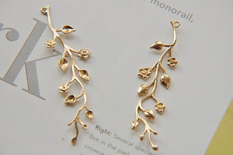 May include: Two gold-tone metal charms shaped like a vine with leaves and flowers. The charms have a loop at the top for attaching to jewellery or other crafts.
