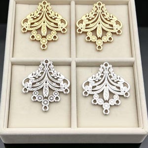 May include: Four metal jewelry connectors arranged in a grid. Two are gold-toned and two are silver-toned. Each piece features a symmetrical, openwork filigree design with leaf-like motifs, geometric patterns, and small circular loops at the base.
