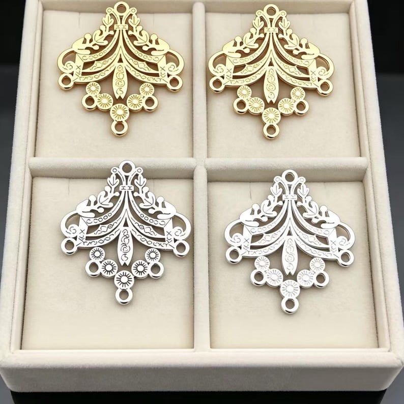 May include: Four metal jewelry connectors arranged in a grid. Two are gold-toned and two are silver-toned. Each piece features a symmetrical, openwork filigree design with leaf-like motifs, geometric patterns, and small circular loops at the base.