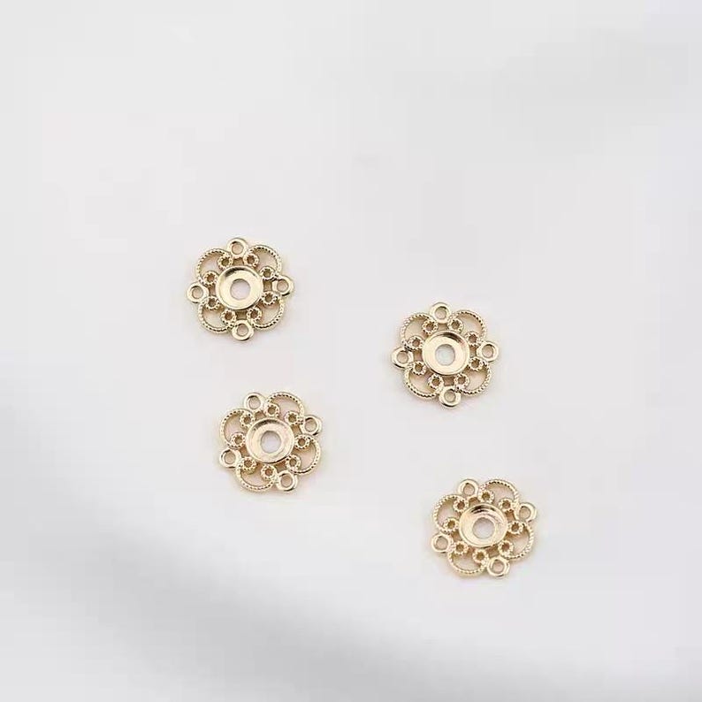 Puede incluir: Cuatro componentes de joyer&iacute;a dorados en forma de flor de filigrana. Cada componente tiene un agujero central rodeado por un dise&ntilde;o calado decorativo con peque&ntilde;os c&iacute;rculos. Los componentes est&aacute;n dispuestos sobre un fondo blanco.