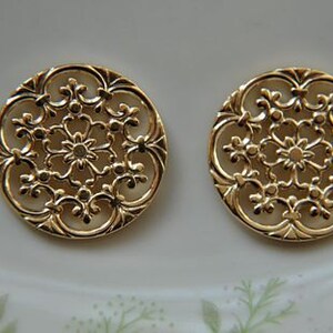 May include: Two gold-toned metal decorative round ornaments with a filigree design. The ornaments have a circular shape with a delicate, intricate pattern of swirls and loops.