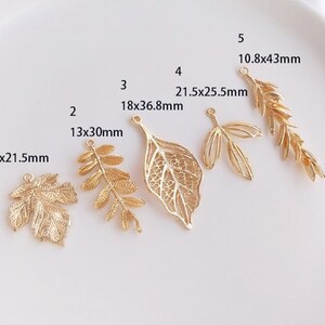 May include: Ten gold leaf charms of various sizes and shapes. The charms are arranged in a row on a white surface. The charms are all made of metal and have a shiny finish. The charms are all different sizes, ranging from 13x30mm to 21.5x25.5mm. The charms are all different shapes, including leaves, branches, and a single leaf with a more intricate design.