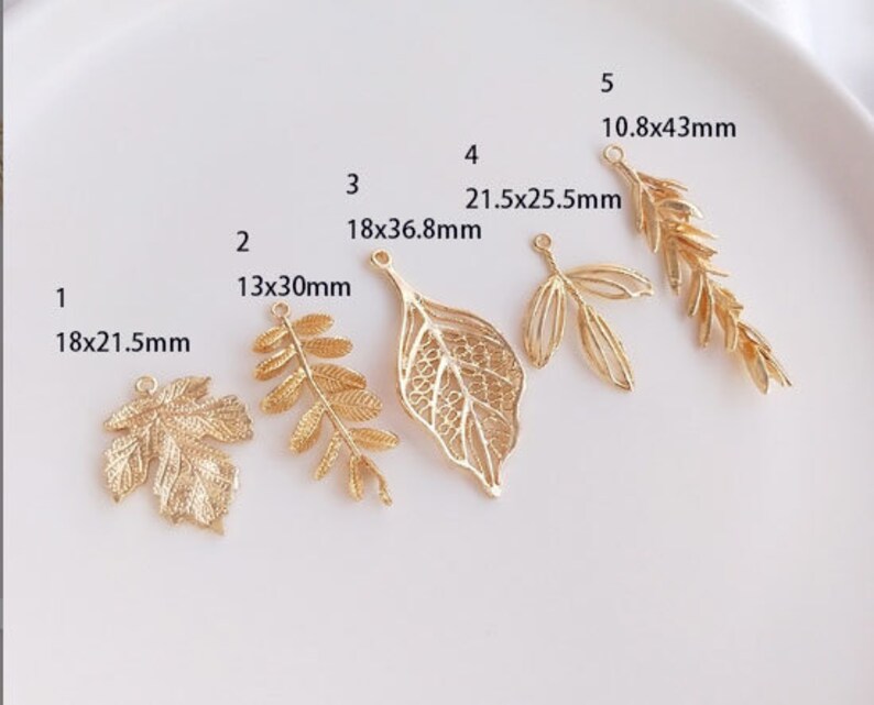 May include: Ten gold leaf charms of various sizes and shapes. The charms are arranged in a row on a white surface. The charms are all made of metal and have a shiny finish. The charms are all different sizes, ranging from 13x30mm to 21.5x25.5mm. The charms are all different shapes, including leaves, branches, and a single leaf with a more intricate design.