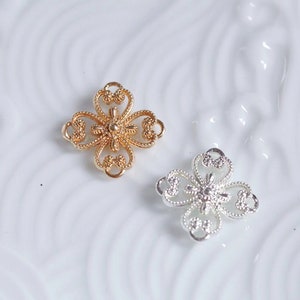 Può includere: Due ciondoli decorativi in metallo a forma di fiore, uno dorato e uno argentato, con disegni intricati e una perla centrale.