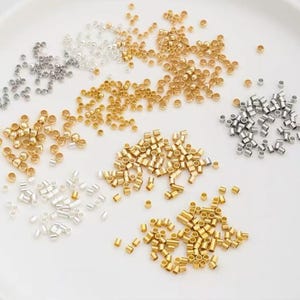 May include: An assortment of small, metallic beads in gold, silver, and bronze tones. The beads are cylindrical and scattered across a white surface, ideal for jewelry making and crafting projects.