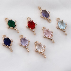 Pode incluir: Berloques de joias douradas com pedras preciosas ovais e coloridas. Os berloques apresentam uma argola na parte superior e uma cauda decorativa, com pequenas pedras de destaque ao redor da gema principal. As cores incluem verde, vermelho, azul, roxo, rosa e transparente.