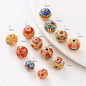 Peut inclure: Onze perles rondes dorées avec des motifs complexes et des accents d'émail colorés. Les perles sont disposées en cercle sur une surface blanche. Chaque perle est numérotée et son diamètre est indiqué en millimètres.