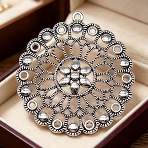 May include: A circular silver-toned metal pendant featuring an intricate openwork filigree design. The center displays a six-petaled flower motif surrounded by concentric layers of loops and small circular accents along the outer scalloped edge.