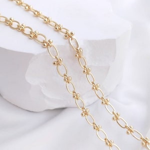May include: Two gold chains with oval links and small round beads. The chains are layered on top of each other and are photographed on a white surface.