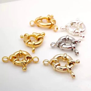 May include: Close-up of six metal jewelry clasps. Three are gold-toned, and three are silver-toned. Each clasp is circular with a hinged opening and small loops for attachment. The clasps are arranged on a white surface.