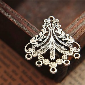 May include: A silver-toned metal jewelry connector featuring an intricate filigree design with symmetrical leaf-like patterns, a central vertical spine, and five small circular loops along the bottom edge for attaching additional components.