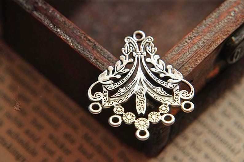 May include: A silver-toned metal jewelry connector featuring an intricate filigree design with symmetrical leaf-like patterns, a central vertical spine, and five small circular loops along the bottom edge for attaching additional components.