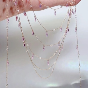 May include: A multi-strand necklace with delicate gold chains. The necklace features small, clear beads and teardrop-shaped pink gemstones. The chains are held by a hand against a soft, neutral background. The jewelry is elegant and suitable for special occasions.