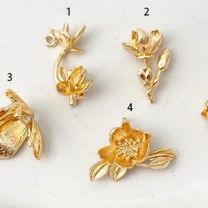 May include: Five gold-coloured floral charms are displayed. The charms feature different flower designs, such as open blooms, buds, and detailed petals. These charms are suitable for jewellery making or craft projects.