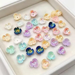 May include: A collection of small, multi-colored flower-shaped beads scattered across a surface. The beads feature various pastel and iridescent finishes, including shades of pink, blue, yellow, purple, and white, each with a central hole.