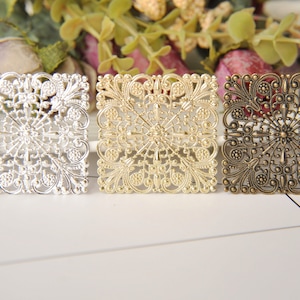 May include: Three square metal embellishments with intricate filigree designs. The embellishments are silver, gold, and bronze.