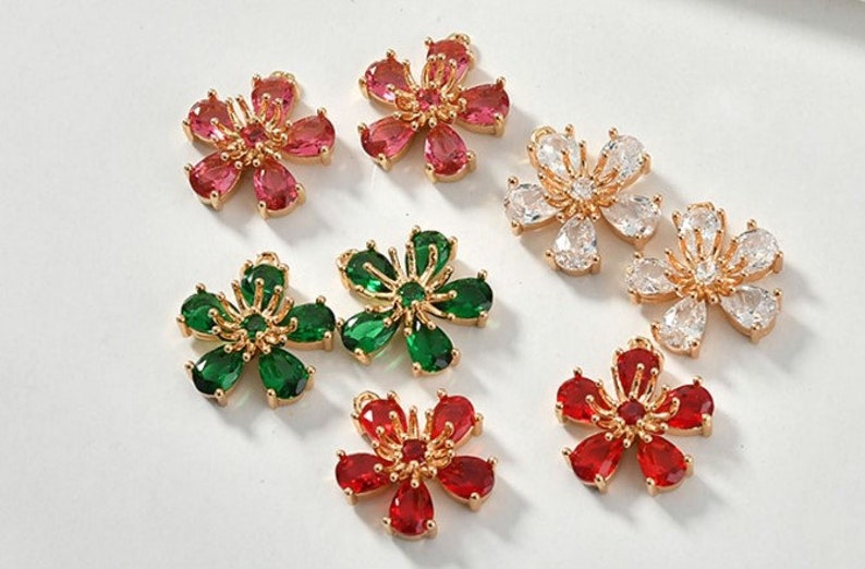 May include: Six gold flower charms with different colored gemstones. The charms are red, green, and clear. The charms are perfect for jewelry making or adding a touch of sparkle to any project.
