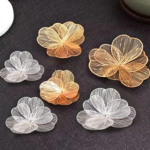 Peut inclure: Un ensemble de six ornements en forme de fleur fabriqués à partir de fines mailles métalliques. Trois ornements présentent une finition dorée brillante, tandis que les trois autres ont une finition argentée, avec des pétales délicats superposés.