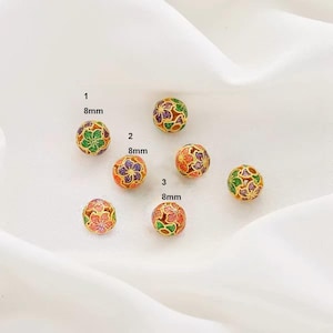 Peut inclure: Six perles dorées de 8 mm avec des motifs floraux colorés. Les perles sont disposées en demi-cercle sur une surface blanche.
