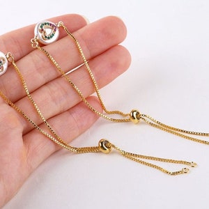 May include: Two gold chain bracelets with adjustable sliding knots. Each bracelet features a white pearl-like charm with a colorful heart design.