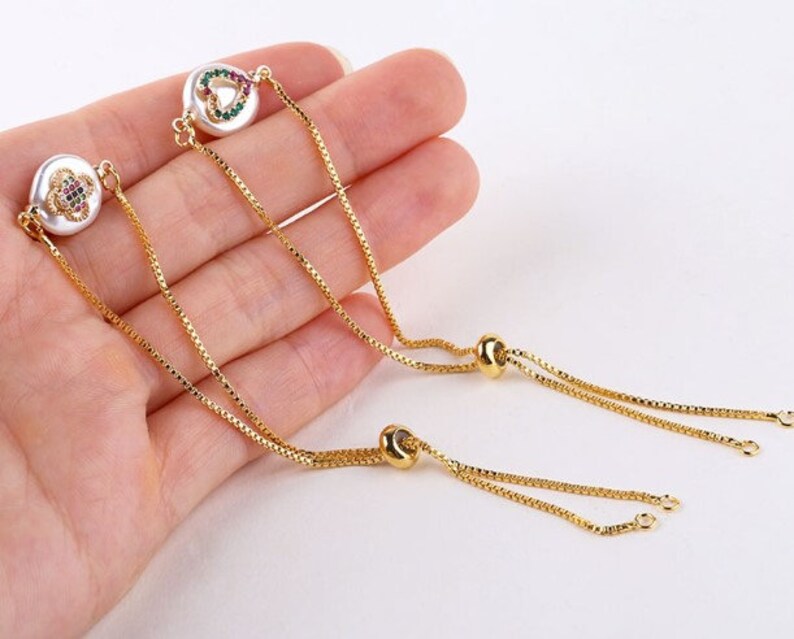 May include: Two gold chain bracelets with adjustable sliding knots. Each bracelet features a white pearl-like charm with a colorful heart design.