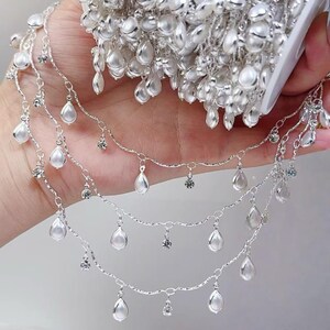 May include: Silver-toned jewelry chain with dangling pear-shaped white beads and small clear crystals. The chain is held in a hand, with a larger quantity of the chain visible in the background.