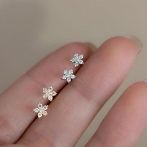 May include: Three pairs of flower-shaped earrings with clear cubic zirconia stones. The earrings are shown on a hand, with the silver earrings on the top, the gold earrings in the middle, and the smaller gold earrings on the bottom.