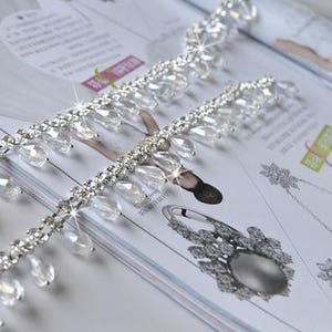 May include: A silver beaded trim with clear teardrop-shaped crystals. The trim is made of small silver beads and clear crystals that are strung together in a repeating pattern. The trim is perfect for adding a touch of sparkle to any project.