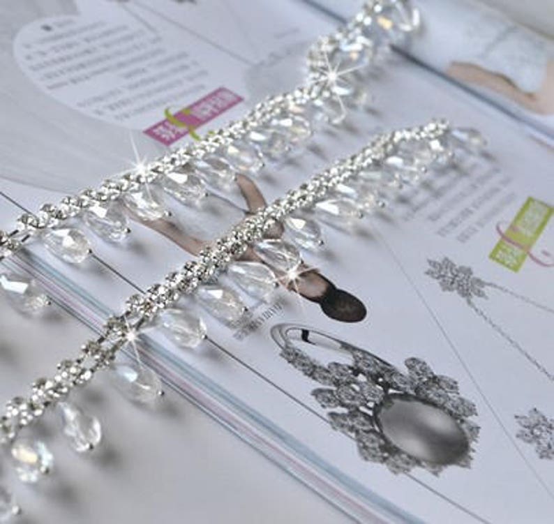 May include: A silver beaded trim with clear teardrop-shaped crystals. The trim is made of small silver beads and clear crystals that are strung together in a repeating pattern. The trim is perfect for adding a touch of sparkle to any project.