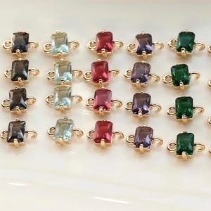 Könnte beinhalten: Eine Auswahl rechteckiger Edelsteinverbinder in verschiedenen Farben, darunter Blau, Grau, Aqua, Rot, Lila, Grün und Klar. Jeder Stein ist in einem goldfarbenen Metallrahmen mit gebogenen Verbindungsösen gefasst.
