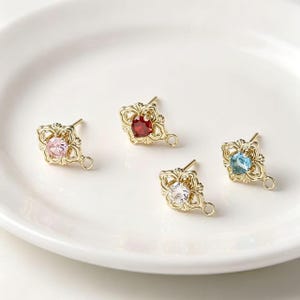 May include: Four gold-toned earring components featuring ornate, filigree-style metal frames. Each piece holds a single round faceted gemstone in the center, with colors including light pink, deep red, clear white, and light blue, plus a small loop.