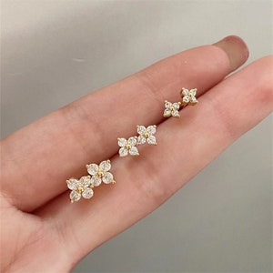 Peut inclure: Trois paires de boucles d'oreilles en or avec un motif de fleur serti de pierres de zircone cubique transparentes. Les boucles d'oreilles sont présentées sur la main d'une personne.