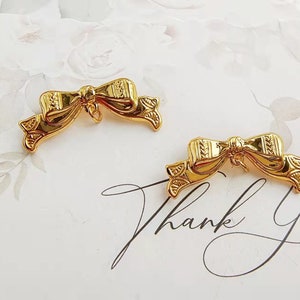 Pode incluir: Dois ornamentos dourados em forma de laço com detalhes intrincados. Os laços estão sobre um fundo branco com desenhos florais e a inscrição manuscrita "Thank You".