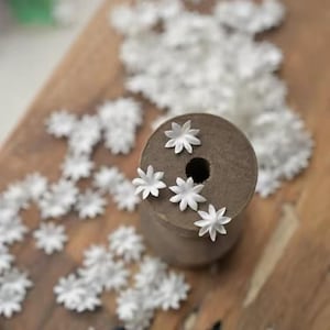 May include: A collection of small, white, star-shaped paper flowers scattered on a wooden surface. Several flowers are attached to a brown spool. The flowers have multiple petals and a delicate appearance, suitable for crafting or decorative purposes.