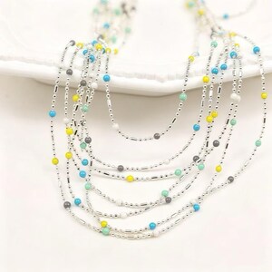 May include: A multi-strand necklace featuring thin silver-toned ball chains interspersed with small, smooth beads in vibrant shades of turquoise, bright yellow, white, and dark gray, creating a layered and delicate appearance with varied bead placement.