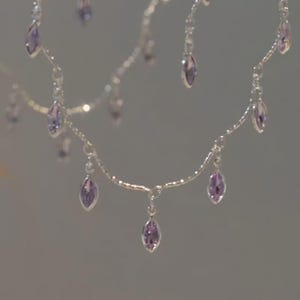 Könnte beinhalten: Eine silberfarbene Kettenhalskette mit mehreren herabhängenden, marquise-geschliffenen lavendelfarbenen Edelsteinen. Die feine Metallkette ist bogenförmig drapiert, wobei jeder einzelne violette Stein in einer kleinen silberfarbenen Metallfassung sitzt.