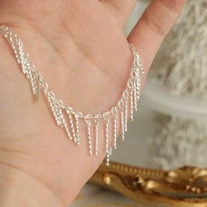 May include: A silver-toned chain necklace featuring a series of short, dangling, twisted bar charms spaced evenly along the length of the chain. The metallic links are connected in a delicate pattern, creating a fringe-like effect across the design.