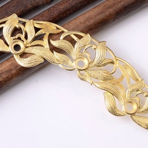 Puede incluir: Un componente metálico calado dorado con un diseño floral. La pieza presenta hojas caladas y engastes circulares, posiblemente para adornos. La forma curva sugiere su uso como elemento decorativo en joyería o artesanía.