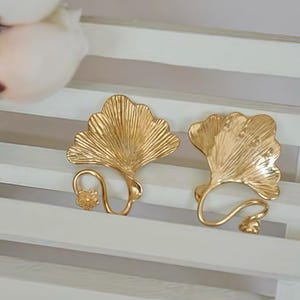 May include: Two gold-colored metal ginkgo leaf-shaped decorative objects. Each leaf has detailed veining and is attached to a curved stem with a small flower detail. The objects are displayed on a white slatted surface.