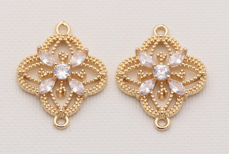 May include: Two gold-tone metal charms with a decorative, openwork design featuring a central clear rhinestone and smaller rhinestones around the edges. Each charm has two loops for attaching to jewelry.