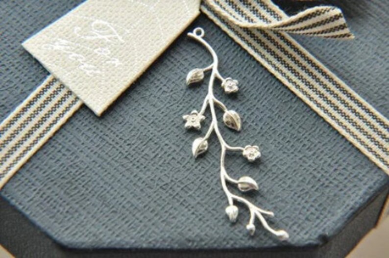 May include: Silver pendant featuring a delicate branch with leaves and flowers. The pendant is on a dark grey background with a white and black striped ribbon.