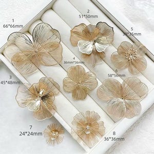 May include: A collection of eight gold-colored, wire-frame flower embellishments of varying sizes, from 24mm to 66mm. The flowers are arranged in a white display tray, showcasing their delicate, openwork design. Each flower has a unique petal arrangement.