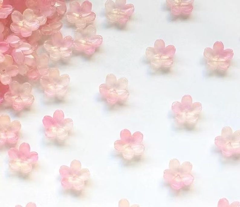 May include: A collection of translucent, pink flower-shaped beads scattered on a white surface. The beads have a delicate, iridescent quality, with varying shades of pink and white, creating a soft, ethereal appearance. Ideal for crafting and jewelry making.