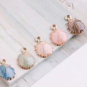 May include: Five seashell charms in a row, each with a gold-coloured loop at the top. The shells are in shades of blue, green, pink, and brown, with a glittery finish. The charms are on a white and grey surface.
