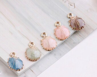 May include: Five seashell charms in a row, each with a gold-colored loop at the top. The shells are in shades of blue, green, pink, and brown, with a glittery finish. The charms are on a white and gray surface.